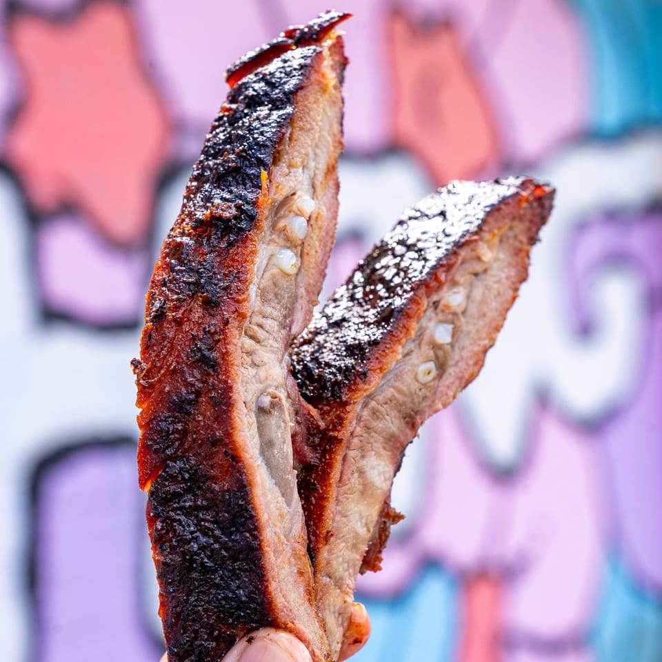 Two Supreme Barbecue ribs, charred and glistening, held against a colorful, blurred background.