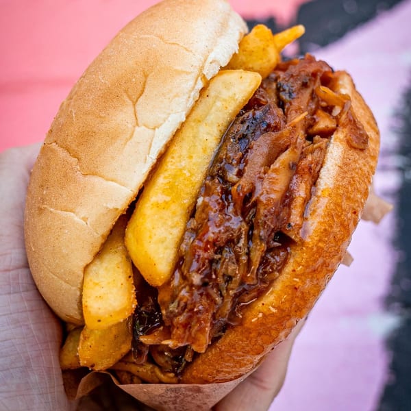 Supreme Barbecue pulled pork sandwich with fries, held in a hand against a pink background.