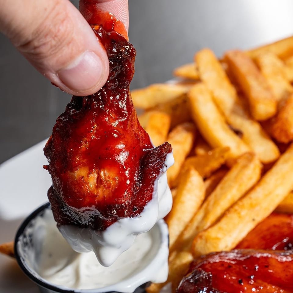 Hand dipping a Supreme Barbecue chicken wing into a white sauce, with fries in the background.