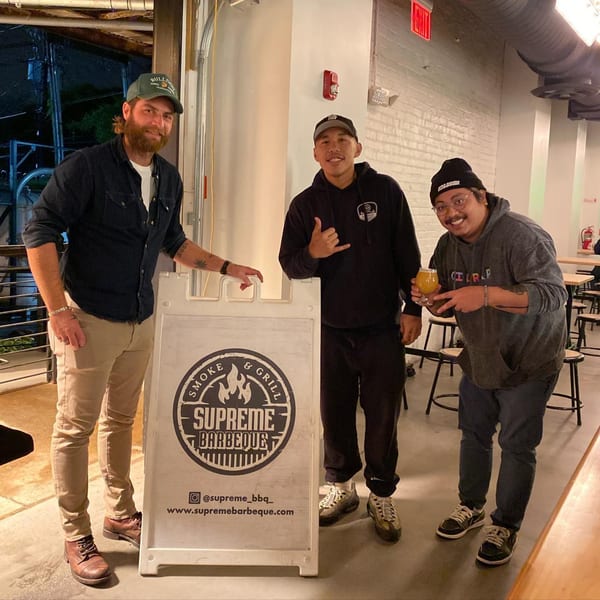 Three men smiling beside a Supreme Barbecue sign in a restaurant setting.