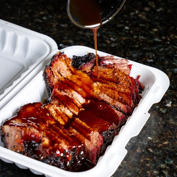 BBQ sauce being drizzled from a ladle onto sliced brisket in a white takeout container.