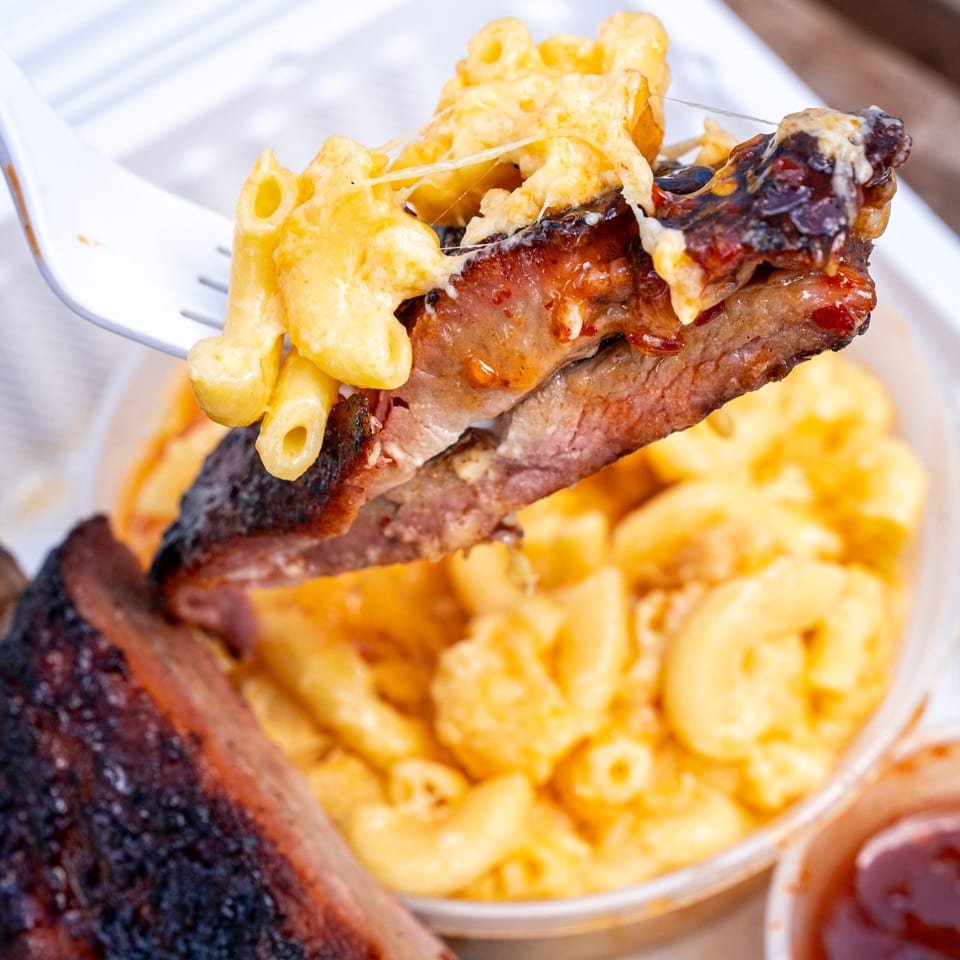 A fork lifts a piece of Supreme Barbecue rib topped with cheesy macaroni, with more mac and cheese in the background.